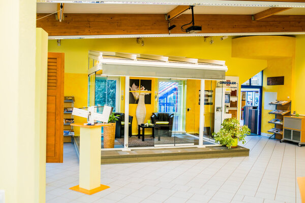 Moderner Ausstellungsraum mit Wintergarten, Pflanzen und Sitzbereich auf Fliesenboden.